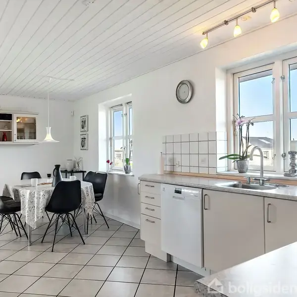 Køkkenbord med vask og opvaskemaskine, omgivet af lysindfald fra vinduer, og spisestue med sort-stole og hvid dug, under en loftlampe. grønne planter ses i baggrunden.
