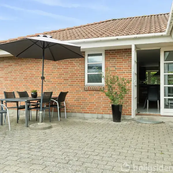 Terrasse med bord og stole under en stor parasol, placeret foran et murstenshus med en havedør åben til et rum indenfor. En plante i krukke står ved døren.