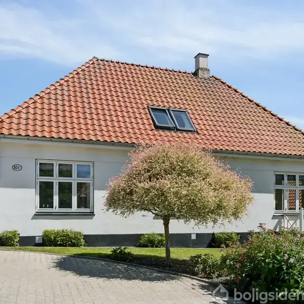 Hus med rødt tegltag og hvid facade, har en forhave med en lille træ og blomster. Det står i en grøn, landlig omgivelser. Husnummer 29 vises ved indgangen.