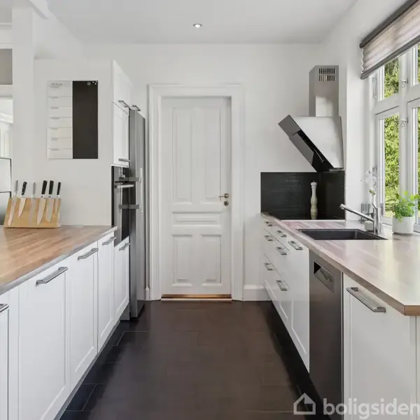 Et moderne køkken med hvide skabe og træbordplader. En håndvask og komfur er placeret ved vinduer, der giver udsigt til grønne træer udenfor. Knivblok og indbyggede ovne ved døren.