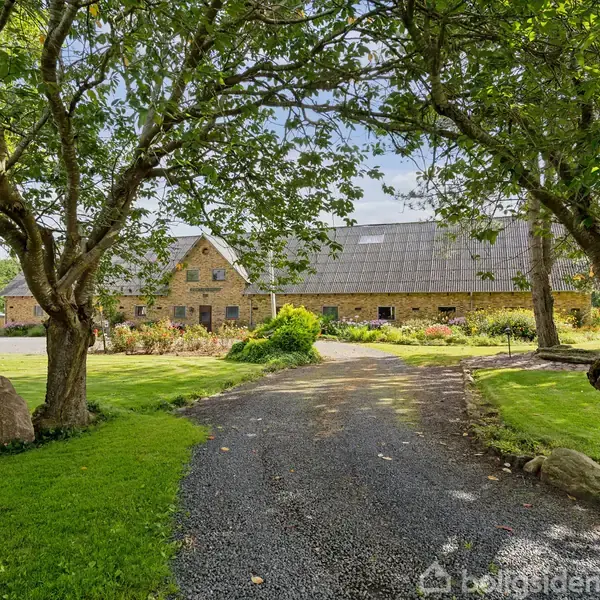Stenbygning står i baggrunden, omgivet af frodig have. En grusvej fører gennem træer og grønt græs til bygningen. Sollys filtrerer gennem løvet, der skaber en fredelig atmosfære.