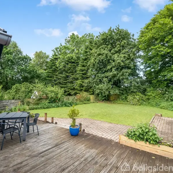Terrasse med træbord og stole står på en trædæk i en have. Omkring er græsplæne, grønne buske og træer under en blå himmel.