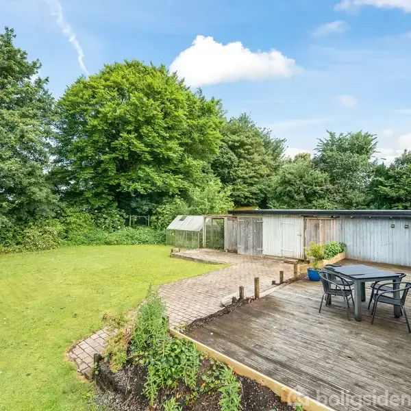 Terrasse med havemøbler står foran et græsareal omgivet af tætte træer. Et skur ses i baggrunden, og himlen er blå med få skyer.