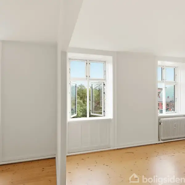 Et lyst værelse med trægulv har tre store vinduer åbne mod udsigt til træer og himmel. Det er delt af en hvid væg, og radiatorer er under vinduerne.