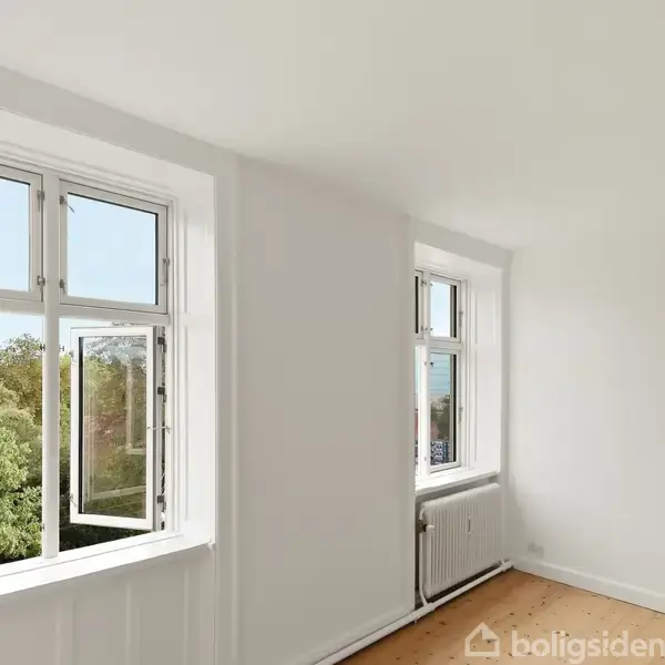 Et lyst rum med hvide vægge har to vinduer åbne mod en udsigt af grønne træer. Rummet har trægulve og en radiator under det mindre vindue.
