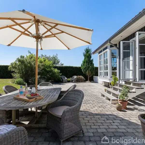 Et gråt parcelhus med rødt tag ligger i en velholdt have omkranset af hække. En carport ses ved siden af huset, som ligger i et roligt villakvarter med grønne områder.