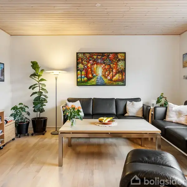 Stue med sort sofa og lænestole står rundt om et trækaffebord med blomster og frugtskål. Vægbillede af farverig skovscene hænger. Lamper og grønne planter ved siden.