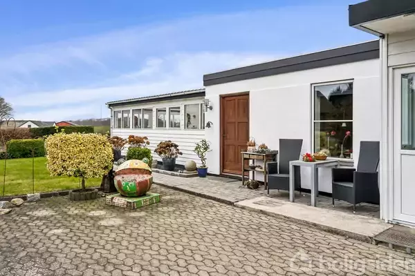 En hvid bygning med en brun dør, foran står to sorte stole og et bord. Omkring er der en flisebelagt gårdsplads og velplejede buske.
