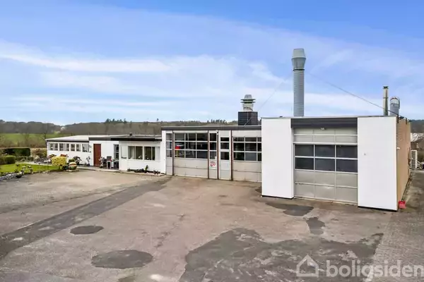 En industriel bygning står stille på en asfalteret plads med store garageporte og rækker af vinduer; baggrunden omfatter grønne træer og en blå himmel.