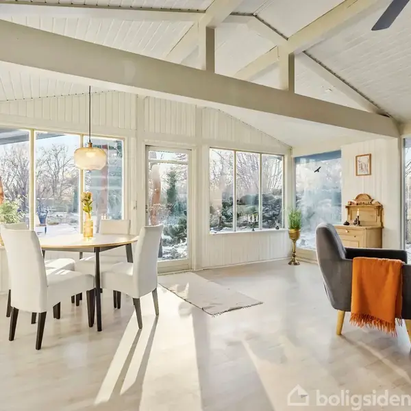 Et lysekronebelyst spisebord med hvide stole står i et lyst rum med store vinduer, der giver udsigt til en snedækket have. En grå lænestol står med et orange tæppe.