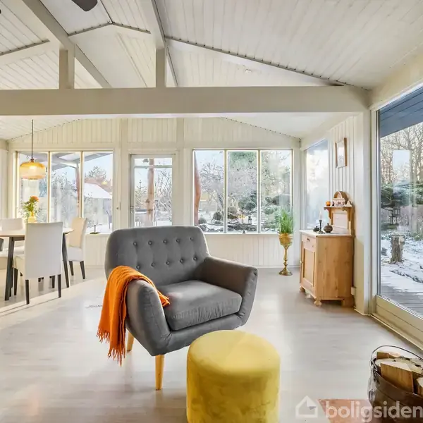 Grå lænestol med orange tæppe står foran gul puf i stue med store vinduer. Spisebord med stole til venstre, brændeovn med ild til højre, udenfor snedækket have.