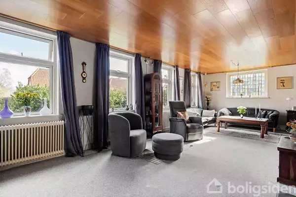 Stue med grå møbler inkluderer to lænestole, en sofa og et sofabord. Store vinduer med mørke gardiner giver naturligt lys. Træloft og tæppebelagt gulv skaber en hyggelig atmosfære.