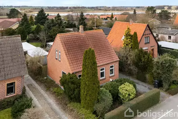 Rødt murstenshus med brunt tegltag, omgivet af velplejede buske og træer, placeret i en række af lignende huse i et grønt nabolag med udsigt til åbent land i baggrunden.