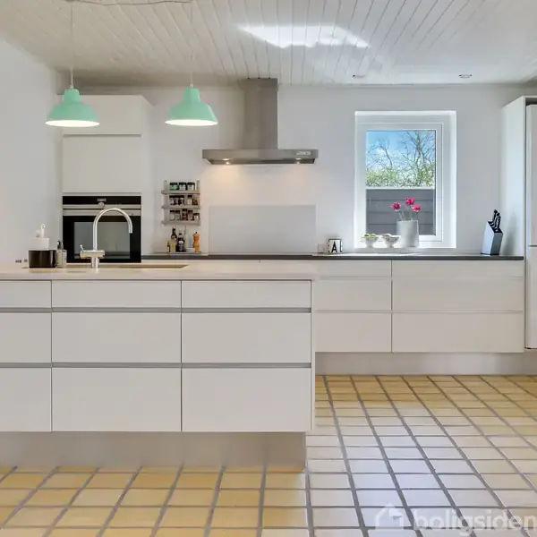 Et moderne køkken med hvide skabe og en stor central ø. To grønne loftslamper hænger over øen. I baggrunden ses et vindue, ovn, og køleskab.