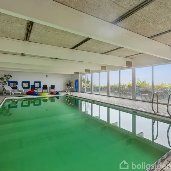 Indendørs swimmingpool med grønligt vand omgivet af fliser. Langs væggen står hvide liggestole og nogle dekorationer. Store vinduer i højre side giver udsigt til grønne omgivelser udenfor.