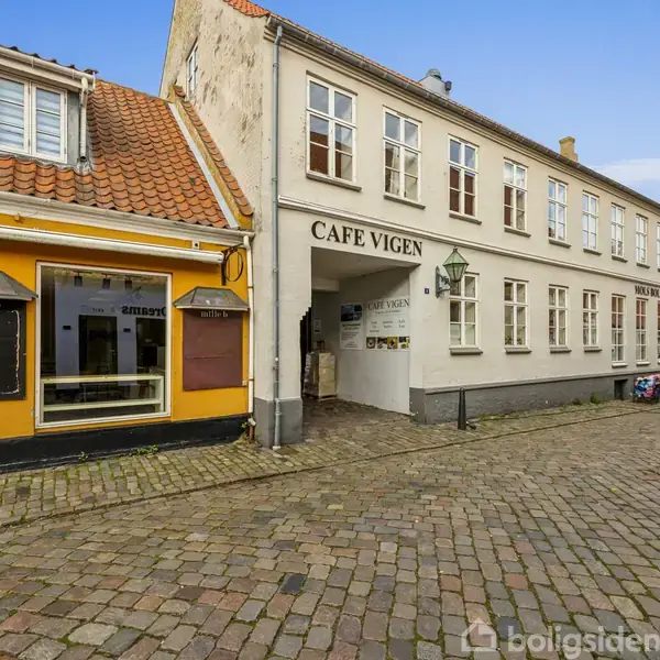En hvid bygning med skiltet "CAFE VIGEN" i en brolagt gade. Til venstre en gul bygning med butiksvindue. Personer i baggrunden går langs gaden.