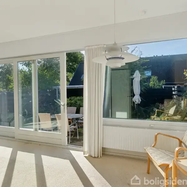 En lys stue med en gyngestol placeret ved vinduet. Store glasdøre og vinduer lader sollys skinne ind, og udenfor ses havemøbler og en parasol på terrassen.