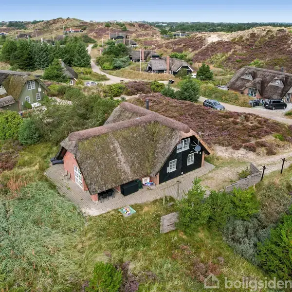 Stråtækt hus står stille i et naturskønt landskab med grønne marker og buske omkring. Et par andre lignende huse ses i baggrunden blandt let kuperet terræn.