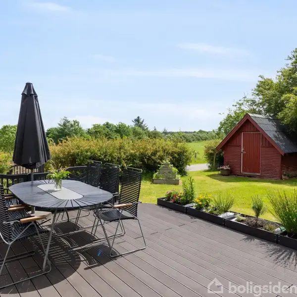 En terrasse med et rundt bord og seks stole, overdækket af en parasol, står foran en grøn have med et rødt skur i baggrunden.
