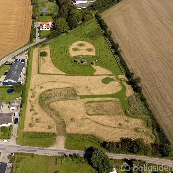 Lufthaven præsenterer en mark formet som en smiley-figur med vej og stier. Omkringliggende huse og marker skaber en landlig kontekst.