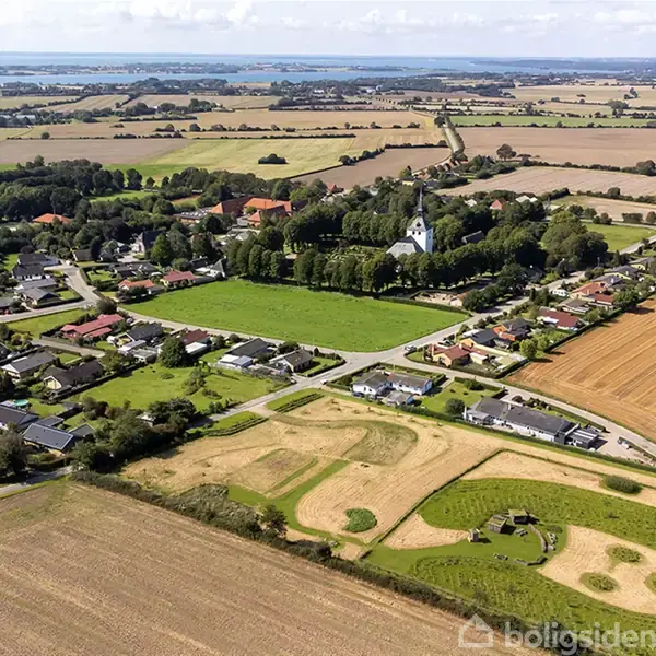 Luftfoto af en landsby med huse omgivet af marker og en kirke centralt beliggende blandt træer. Landskabet strækker sig mod horisonten med udsigt over vandet i baggrunden.
