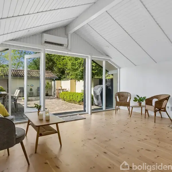 Stue med brændeovn og åbent glasparti, som viser en terrasse med grønne planter. To stole og et lille bord med planter skaber en hyggelig atmosfære.