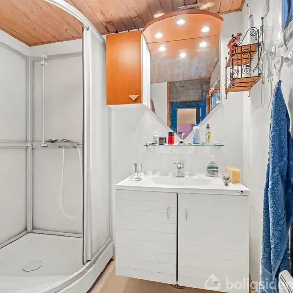 Brusekabine står i hjørnet af badeværelse. Ved siden af er en hvid vask med underskab og spejl. Ovenpå hviler toiletartikler. Rummet er oplyst af indbyggede loftslamper. En blå håndklæde hænger til højre.