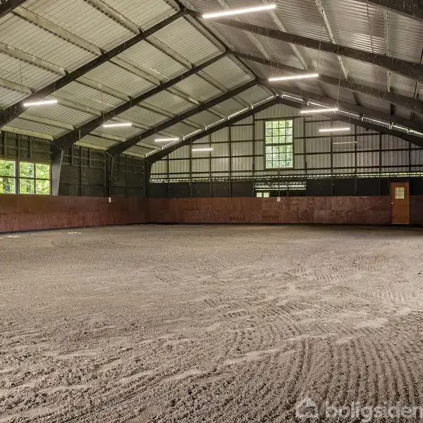 Ridehal med en jordbelagt bund uden heste, oplyst af lange lysstofrør. Vinduer langs væggene skaber naturligt lysindfald, og et trægulv ses langs væggen yderst til højre.