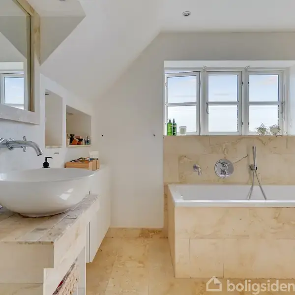 Et lyst badeværelse med en rund håndvask på skabsplads, et rektangulært badekar, og tre små vinduer, placeret i et rum med beige vægge og gulve.