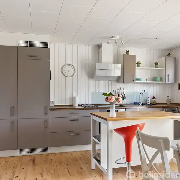 Et lyst køkken med trægulv og hvide vægge. Et køkkenbord har en rød stol og en hvid vase med blomster. Skabe, en emhætte og hylder med planter omringer rummet. En ur hænger på væggen.