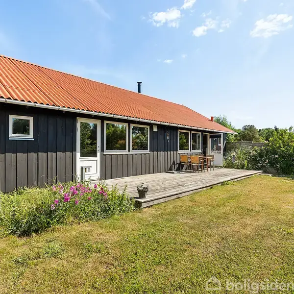 Et sort træhus med rødt tag står stille på en velplejet græsplæne. En terrasse med udemøbler er foran huset, omgivet af grønne buske og klar blå himmel.
