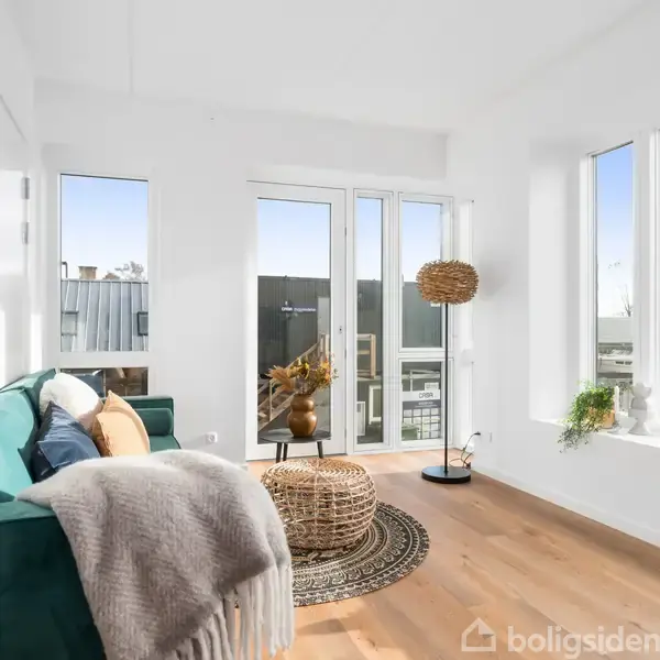 Grøn sofa med tæppe og puder står i et lyst rum med trægulv. En rund fletkurv fungerer som sofabord ved siden af en glasdør, der fører til en balkon.