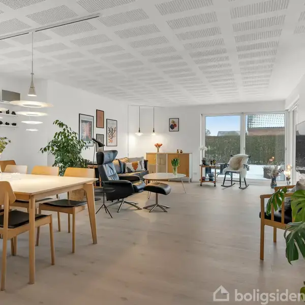 Spisestue med lyst træbord og seks stole, omgivet af lamper, stueplanter og dekorative billeder på væggen. I baggrunden ses en opholdsstue med sofa, stol og stort vindue.