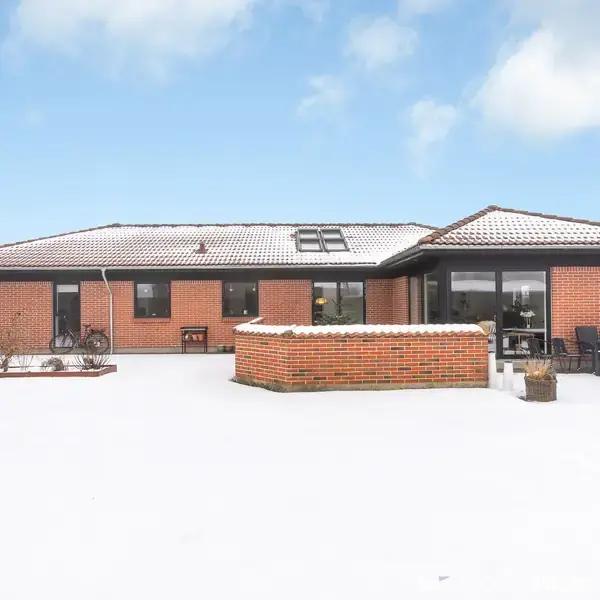 Et murstenshus står stille i en snedækket have, omgivet af en klar himmel. Det har store vinduer og en terrasse med havemøbler til højre.