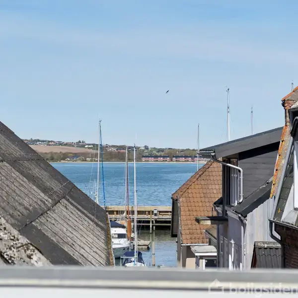 Udsigt over hustage mod en havn, hvor flere sejlmaster er synlige. Baggrunden viser en bugt med en kystlinje i det fjerne under en klar, blå himmel.