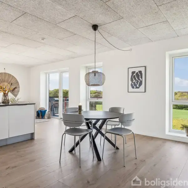 Spisebord med grå stole står i et lyst rum med ovenlys. Køkkenet ligger i baggrunden. Store vinduer giver udsigt over en grøn have. En moderne lampe hænger over bordet.