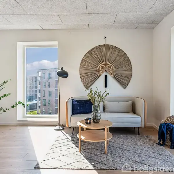 En lys stue med en beige sofa pynret med puder og et blåt tæppe. Foran står et rundt bord med en vase. Væggen har et dekorativt fanformet objekt.