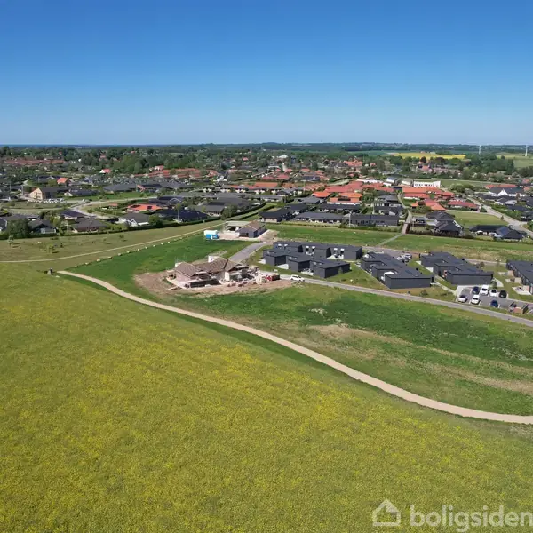 Boligområde under opførelse; flere bygninger ligger i et åbent, grønt landskab. Veje forbinder husene, mens marker og blå himmel udgør den omkringliggende natur.