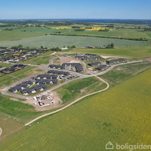 Byområde under opførelse omgivet af åbne marker og veje. Husene er spredt i hjørner af et grønt landskab. Billedet viser en kontrast mellem byggeri og natur.