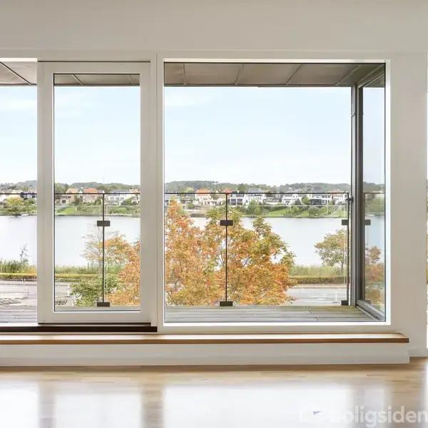 Stort vindue åbent mod balkon, viser udsigt til sø og efterårsfarvede træer i boligområde; lyse trægulve og minimalistisk indretning skaber en lys og åben atmosfære indendørs.