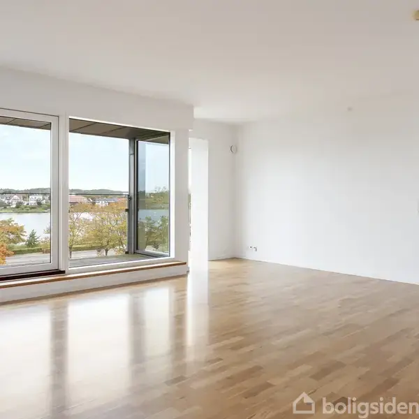 Rummet er lyst med trægulve og store vinduer, der afslører en udsigt over en sø og efterårsfarvede træer. Et tilstødende værelse ses til højre.