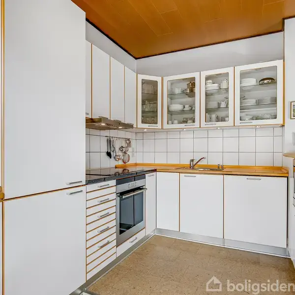 Køkken med hvide skabe, trækanter og indbygget kogeplade. Over vasken er glaslåger, og der hænger en lampe. Rummet er lyst med hvid-flisebeklædte vægge. Tekst: "boligsiden".