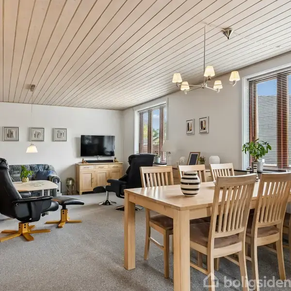 Stue med spisebord og stole placeret til højre under en lysekrone. I baggrunden står en sort lænestol og en sofa foran et TV på en skænk. Vindue til venstre.