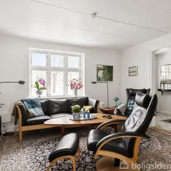 Sort læderstol og sofa, står på et mønstret tæppe, omgivet af lamper og planter ved et vindue. Et sofabord er fyldt med lys og bøger, og rummet er lyst.