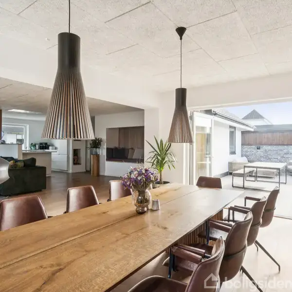 Langt spisebord i træ med blomster i midten omgivet af læderstole. To store pendellamper hænger over bordet. Åben stue med sofa og adgang til terrasse i baggrunden.