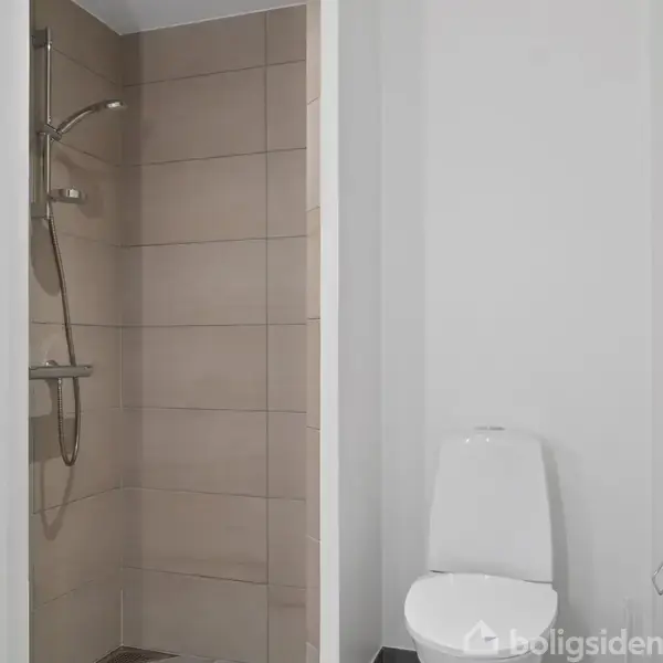 En brusekabine med beige fliser og en bruser i rustfrit stål til venstre. Til højre ses en del af en urinal i et badeværelse.