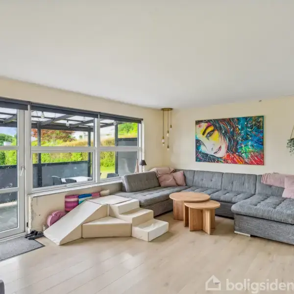Stor, lys stue med grå hjørnesofa og rundt sofabord. Et farverigt maleri hænger på væggen. Store vinduer og glasdør giver udsigt til en have.