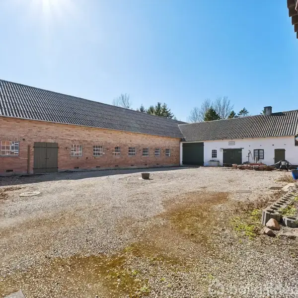 En gårdsplads omgivet af røde murstensbygninger med grønne døre og vinduer. Solen skinner klart på den stenede grund, mens træer i baggrunden antyder en landlig beliggenhed.
