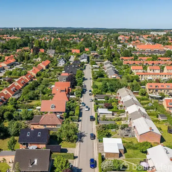 Boligområde set fra luften. Rækkehuse med røde tage ligger langs en vej med parkerede biler. Omgivet af grønne haver og træer, mens byens skyline ses i baggrunden.