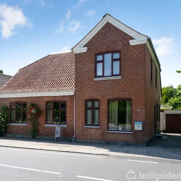 Rødstenshus med skråt tag, står stille ved en vej. Det har store vinduer og en dør på siden. Der er en grussti ved siden af og grønne buske omkring.